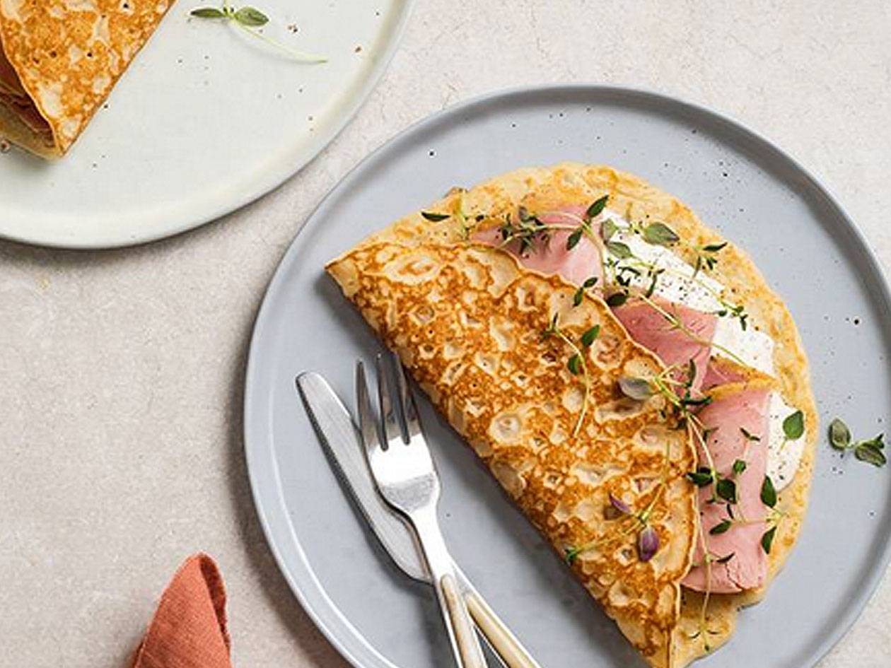 En tallrik med en vikta pannkaka fylld med skinka, färskost och timjan, med en gaffel och kniv bredvid.