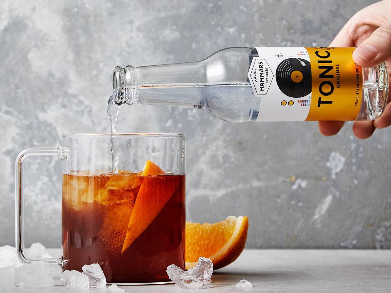 En hand häller Hammar's Tonic i ett glas med en mörk dryck, is och apelsinskiva.