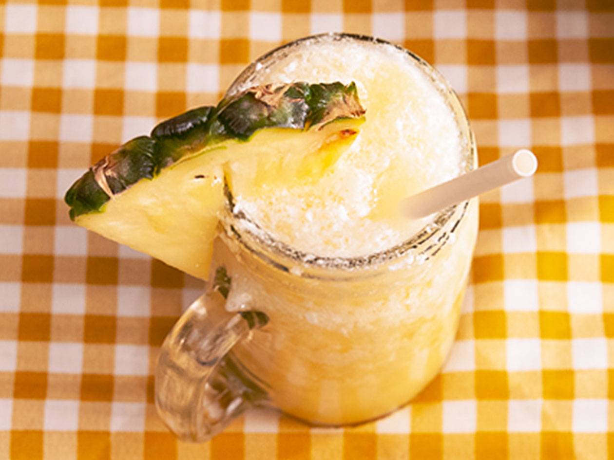 Ananasdrink med sugrör och ananasskiva på en gulrutig duk.