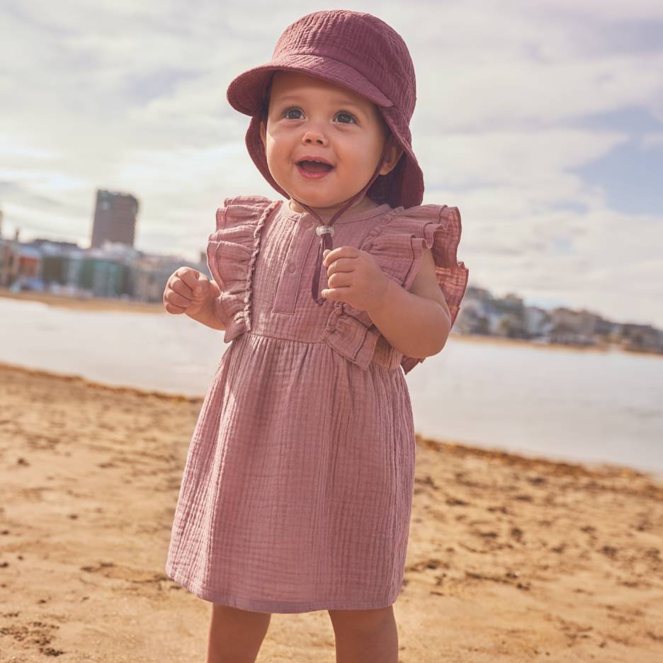 Baby i rosa klänning och hatt på stranden.