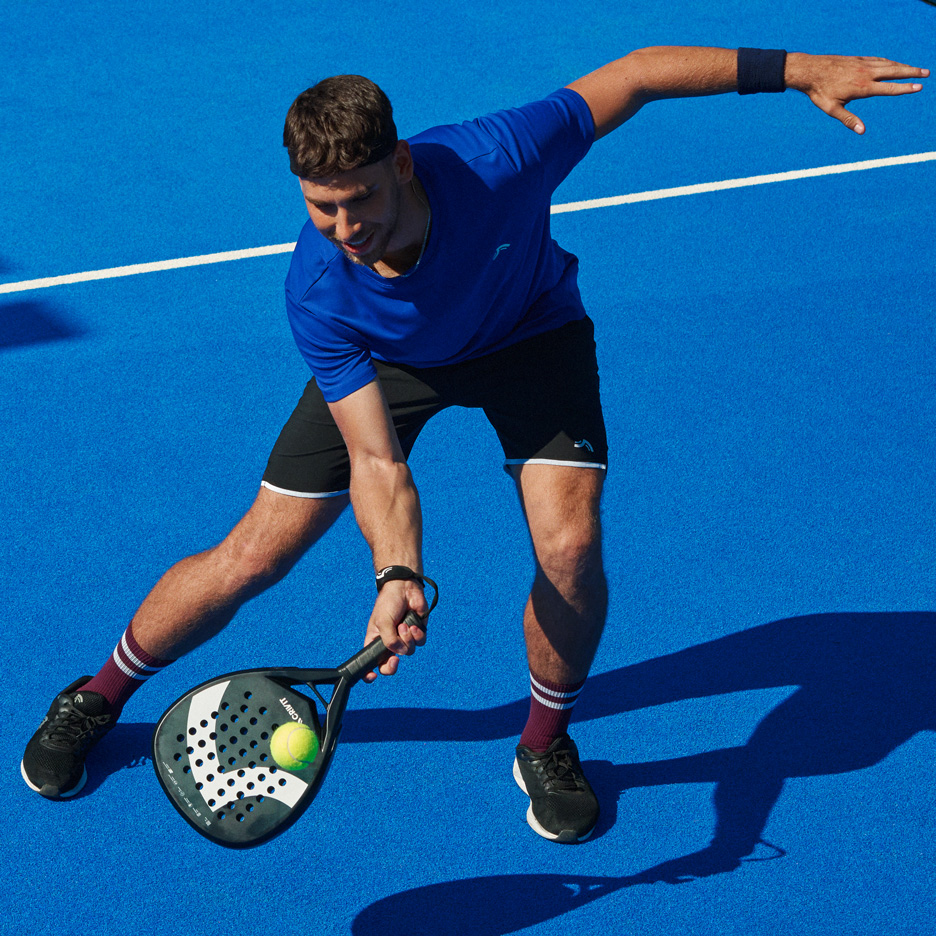 Man i blå t-shirt och svarta shorts spelar padel med racket och boll.