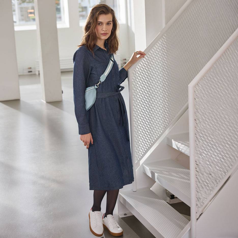 Kvinna i jeansskjortklänning, svarta strumpbyxor och vita sneakers, med en ljusblå midjeväska.