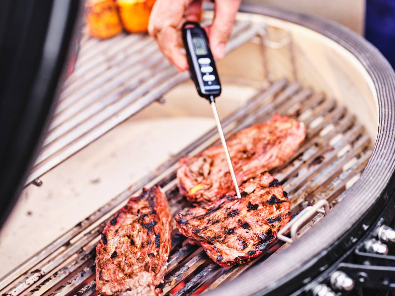 En hand använder en termometer för att kontrollera temperaturen på grillat kött på en grill.