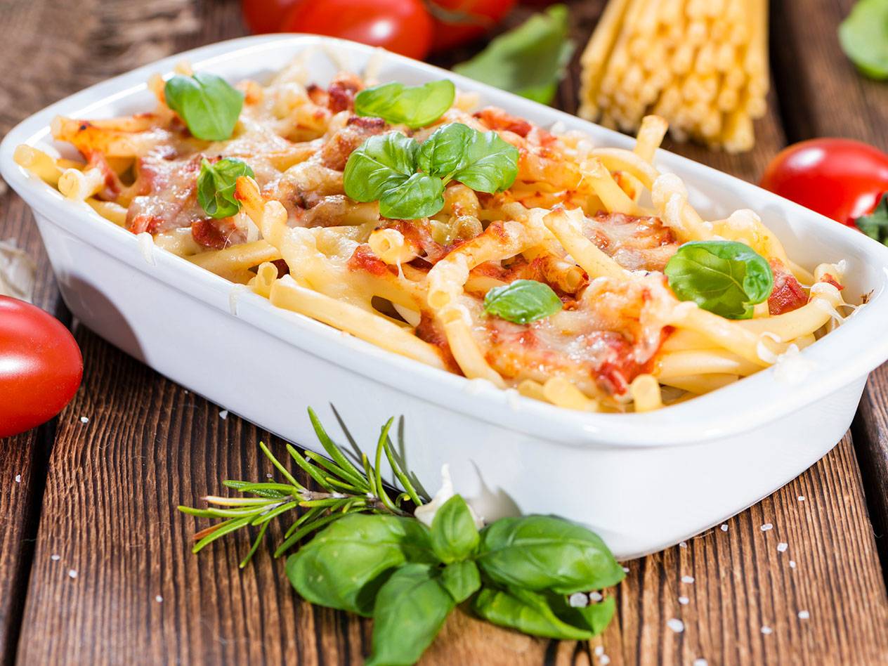 Gratinerad pasta med tomatsås, ost och basilika i en vit form på träbord.