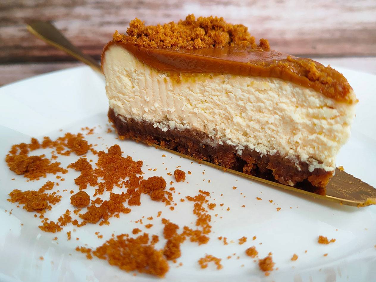 En skiva cheesecake med karamell och smulor på en vit tallrik.
