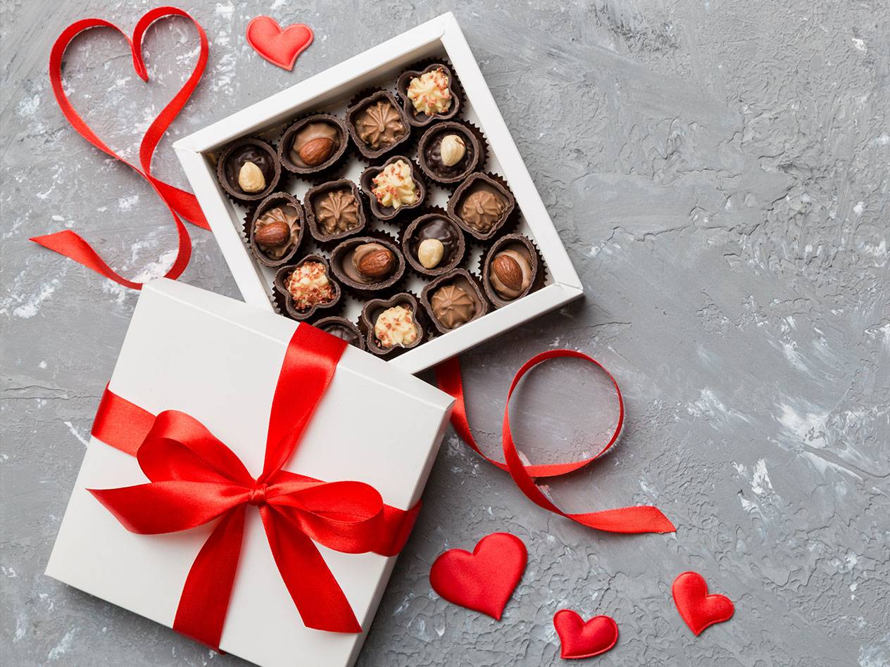 Öppen presentask med chokladpraliner, rött band och hjärtan på grå bakgrund.