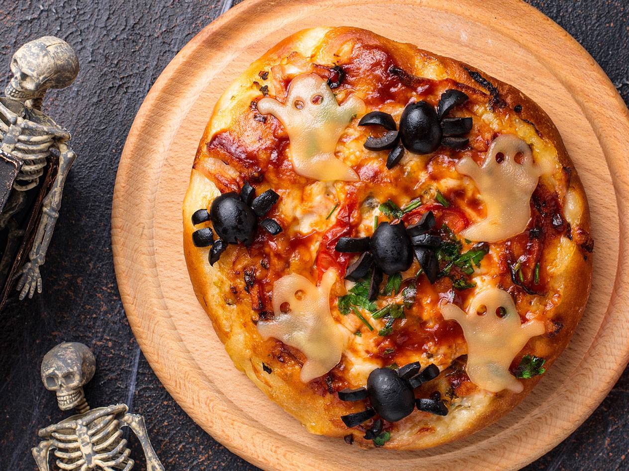 Halloweenpizza med spöken av ost och spindlar av oliver, omgiven av skelettfigurer.