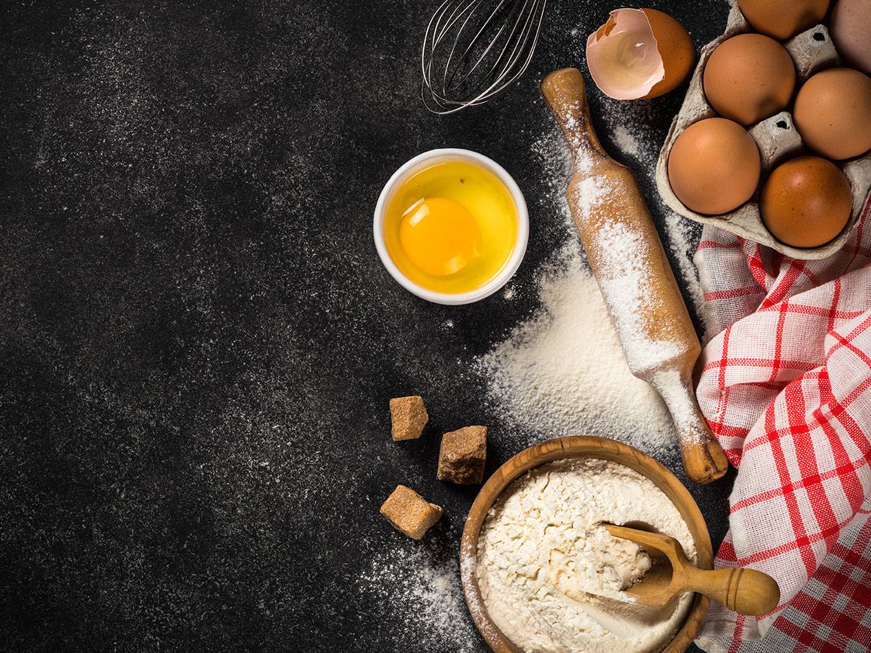 Bakingredienser: ägg, mjöl, kavel och sockerbitar på en mörk bakgrund.
