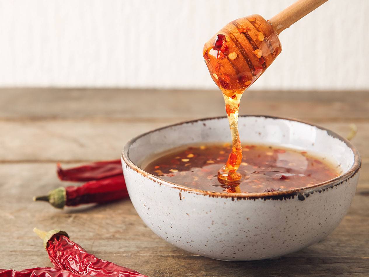 Honung med chili som rinner från en honungsdippare ner i en skål, med torkade röda chilifrukter bredvid.