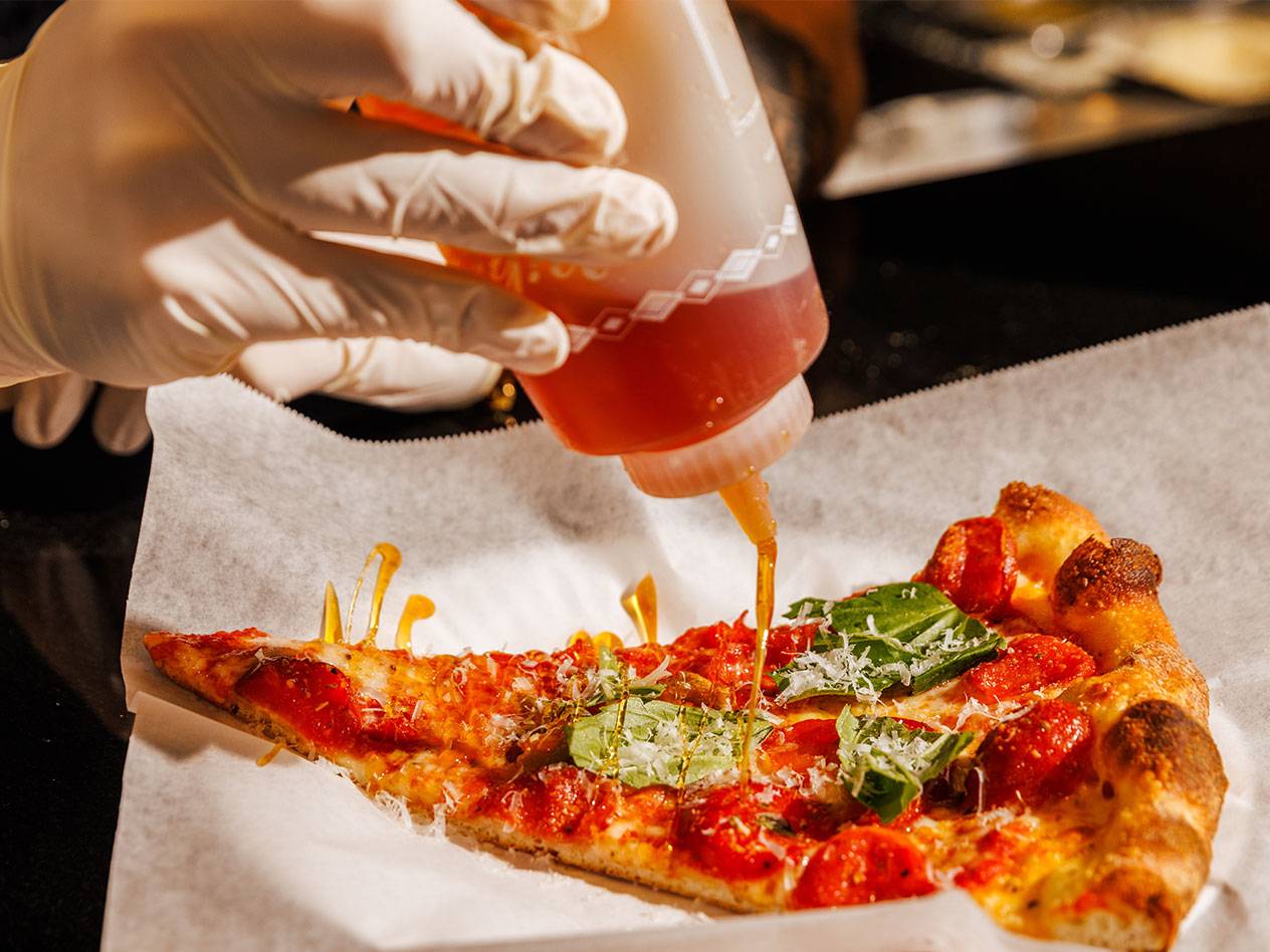 En hand i handske ringlar honung över en pizzabit med tomater, basilika och ost.