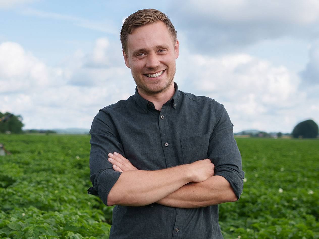 Leende man med korsade armar i ett grönt fält under en molnig himmel.