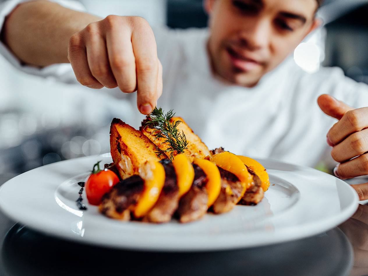 Kock garnerar en maträtt med kött, persikor, potatis och en körsbärstomat.
