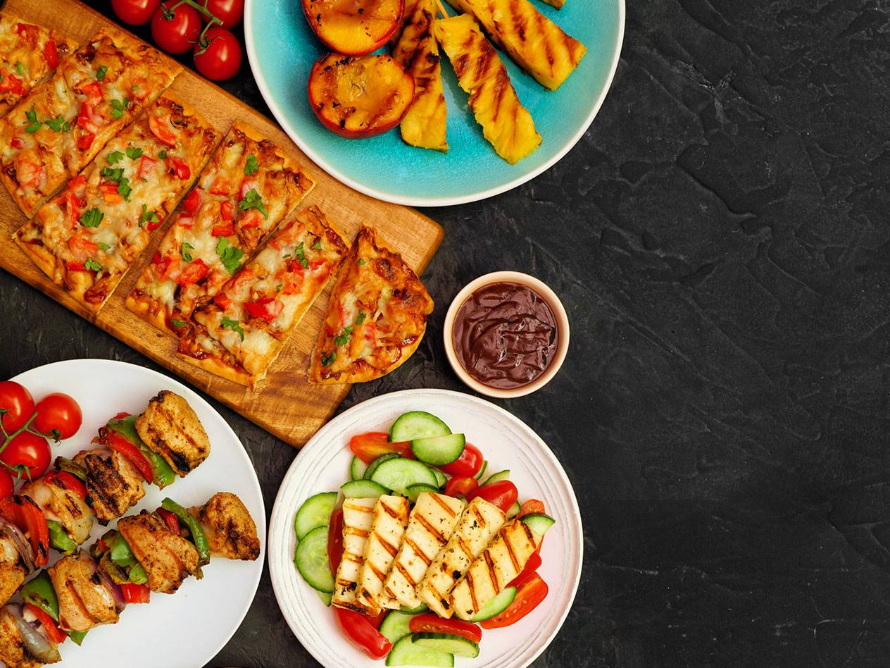 Grillad mat som pizza, kycklingspett, halloumisallad, persikor och ananas på en mörk bakgrund.