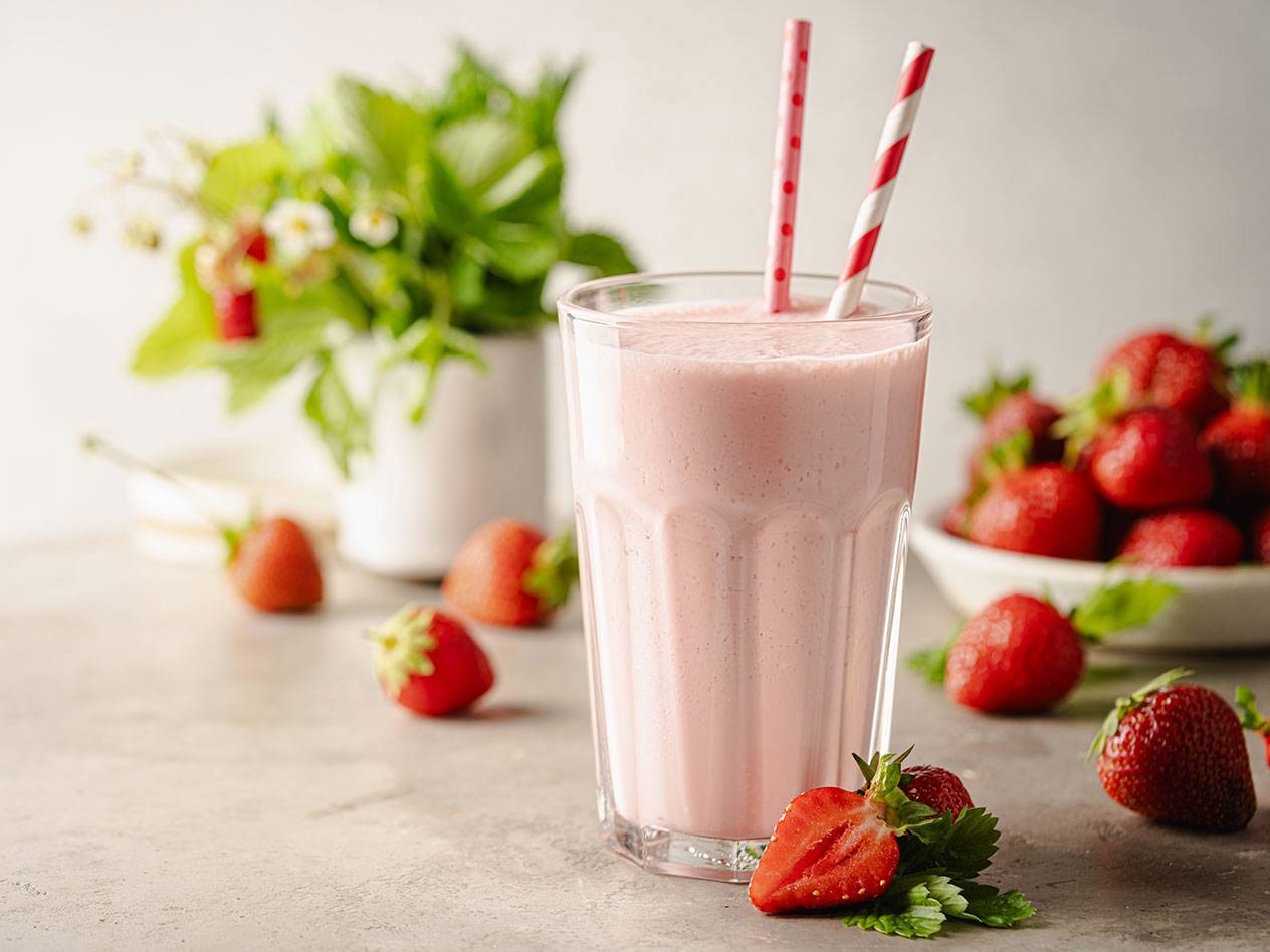 Jordgubbsmilkshake i ett glas med sugrör, omgiven av färska jordgubbar och gröna blad.