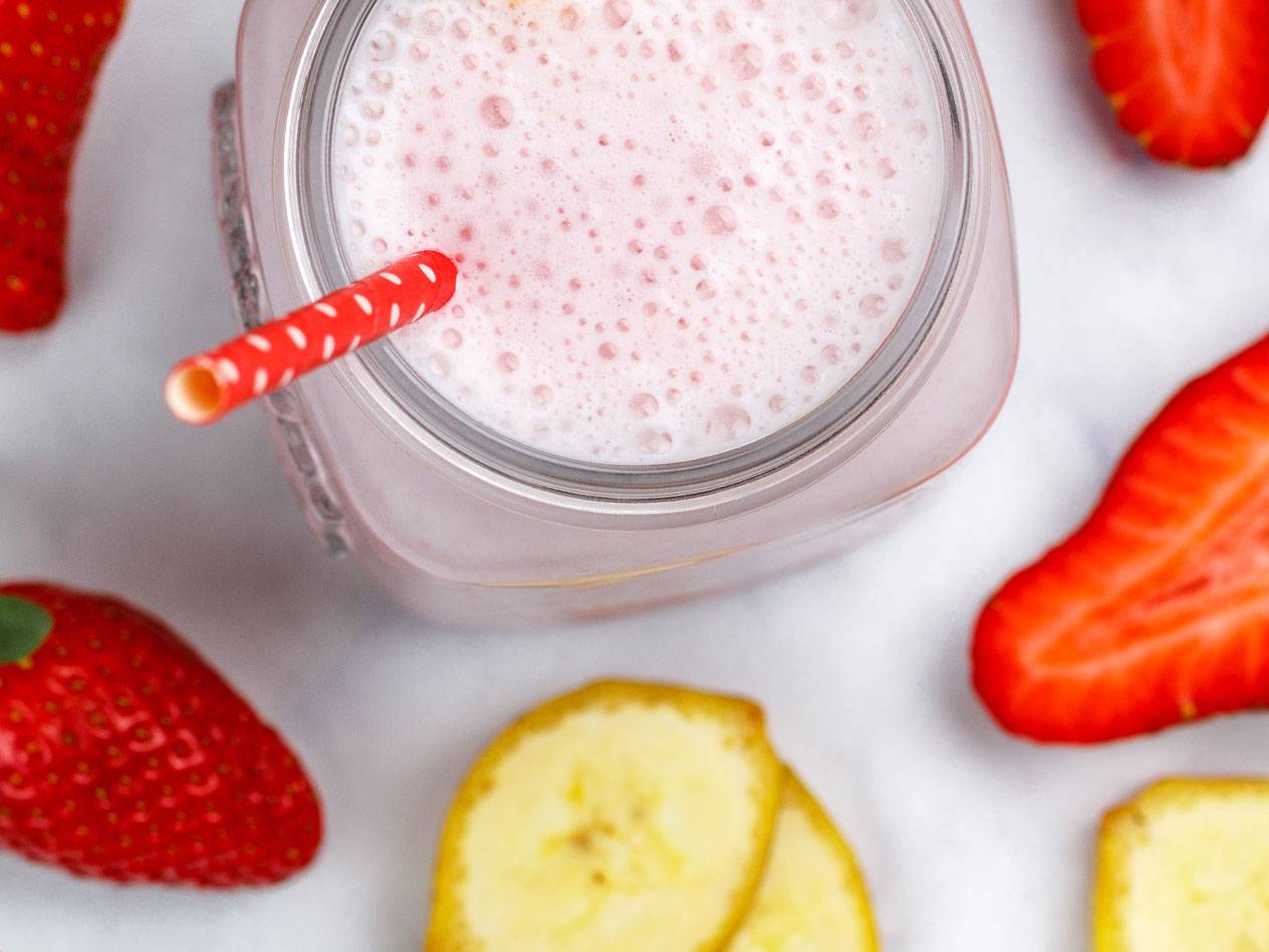 Jordgubbs- och banansmoothie i glasburk med sugrör, omgiven av färska jordgubbar och bananskivor.
