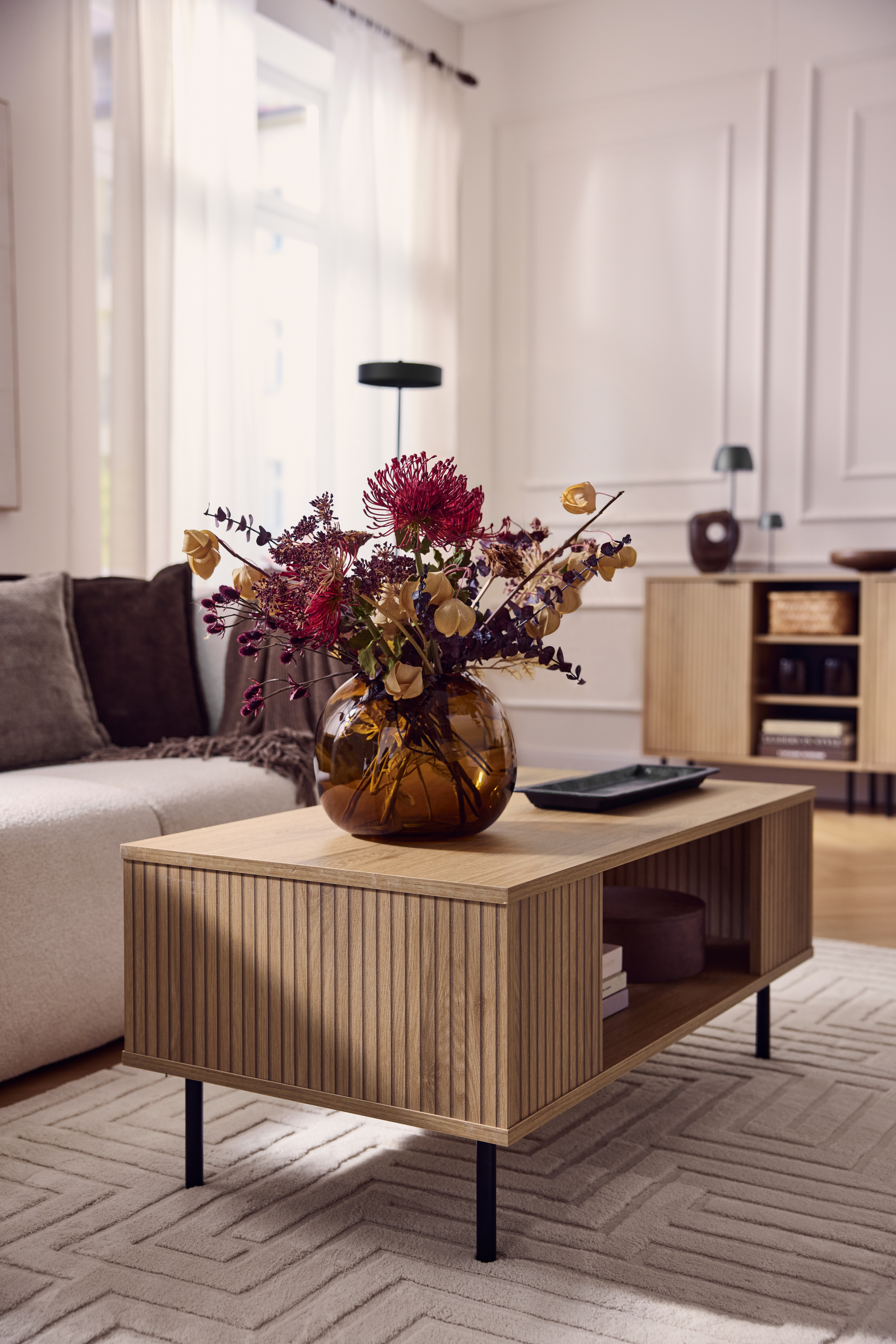 Soffbord i trä med räfflad front, vas med torkade blommor, beige soffa.