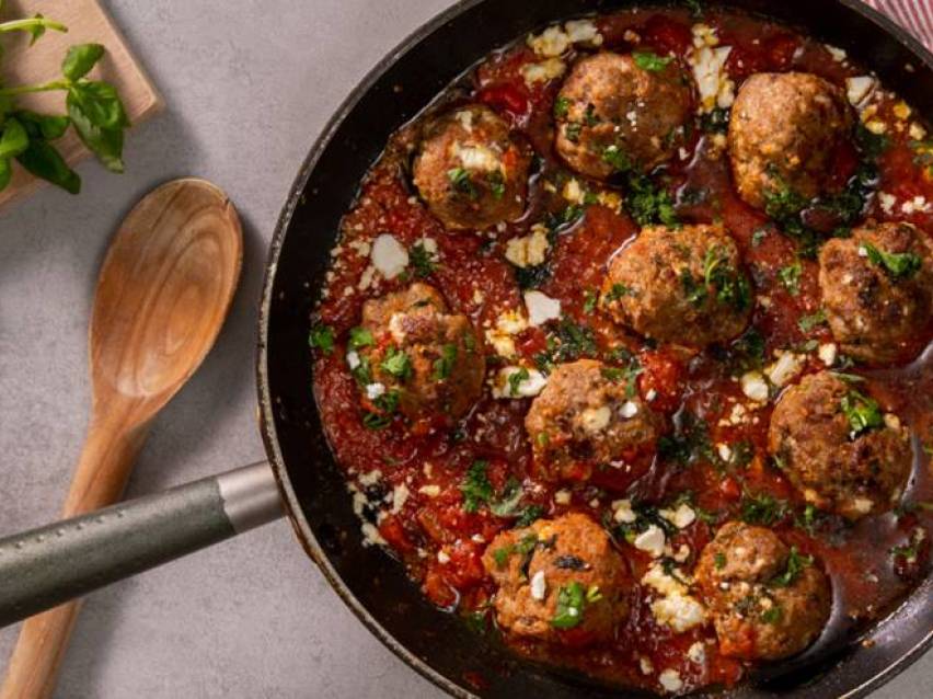 En stekpanna med saftiga köttbullar i en krämig tomatsås, garnerade med fetaost och basilika.
